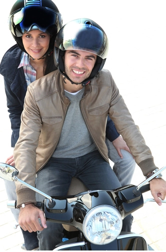 Coppia sorridente in sella a uno scooter a noleggio a Porto Cervo, entrambi con casco protettivo, pronti per esplorare la Sardegna.